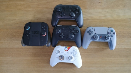 De Switch 2 Pro Controller (midden) vergeleken met: Switch 1 Pro Controller (boven), PS5 controller (rechts), Xbox controller (onder), Joy-Con 2 controller in oplaadstation (links).